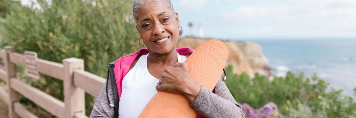 Smiling senior woman holding a yoga mat by the coastline, enjoying wellness and nature.